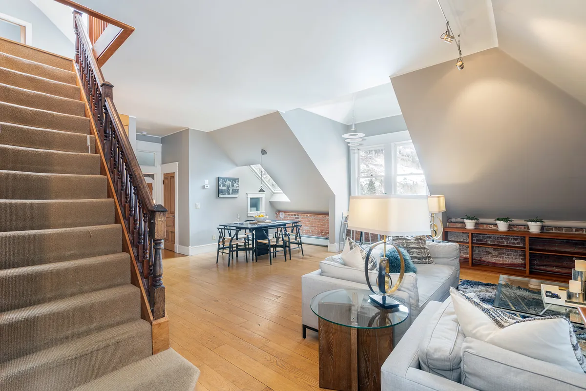 An open-plan view uniting the staircase, living room seating area, and dining table. The space feels airy and connected, featuring cozy furnishings and sunlit windows that reinforce the open-concept layout.