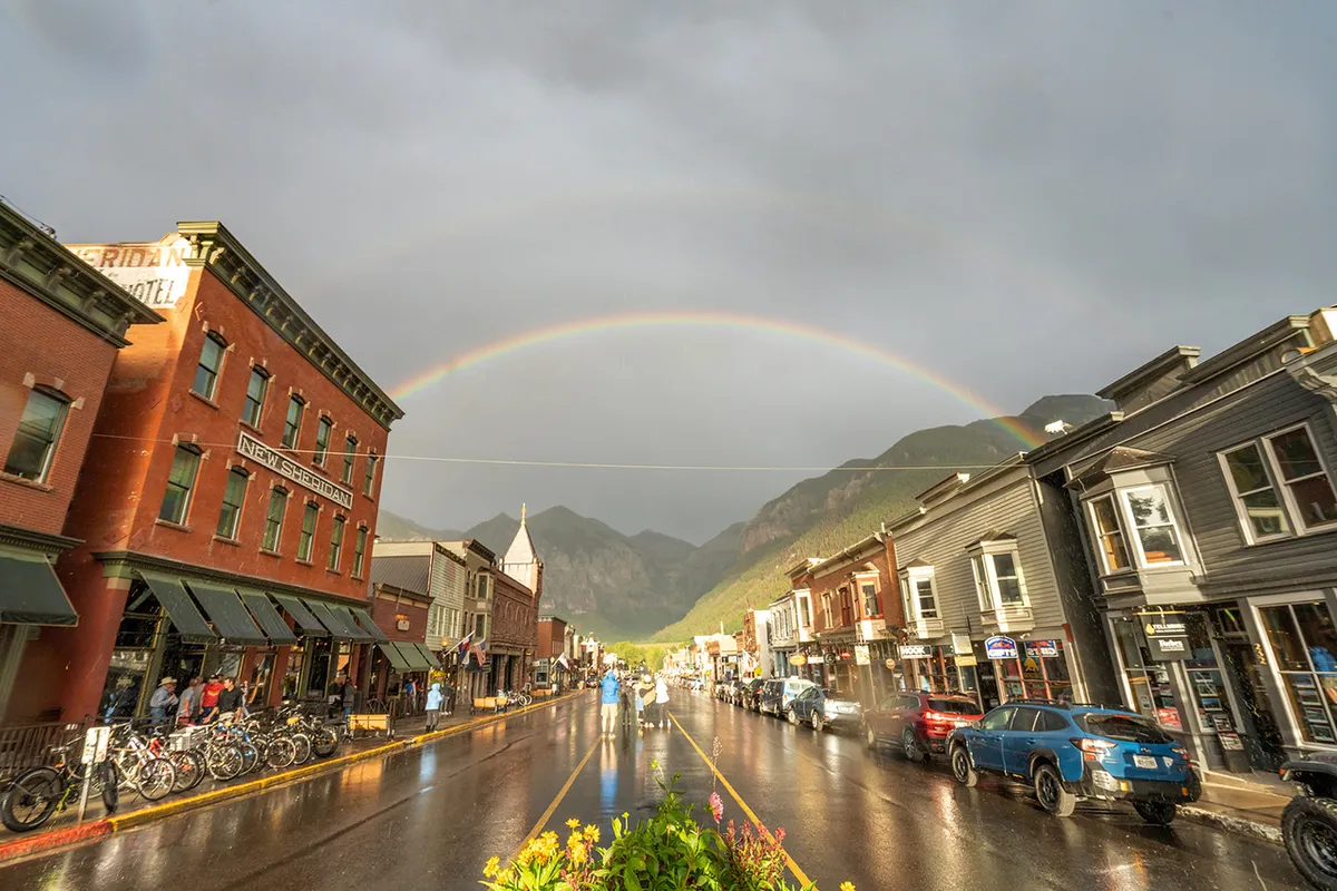A lively street scene features historic buildings, a vibrant rainbow arching overhead, wet pavement from a recent rain, bicycles, and people strolling. The atmosphere is optimistic and bustling, with visible signs of dining, shopping, and cultural exploration.