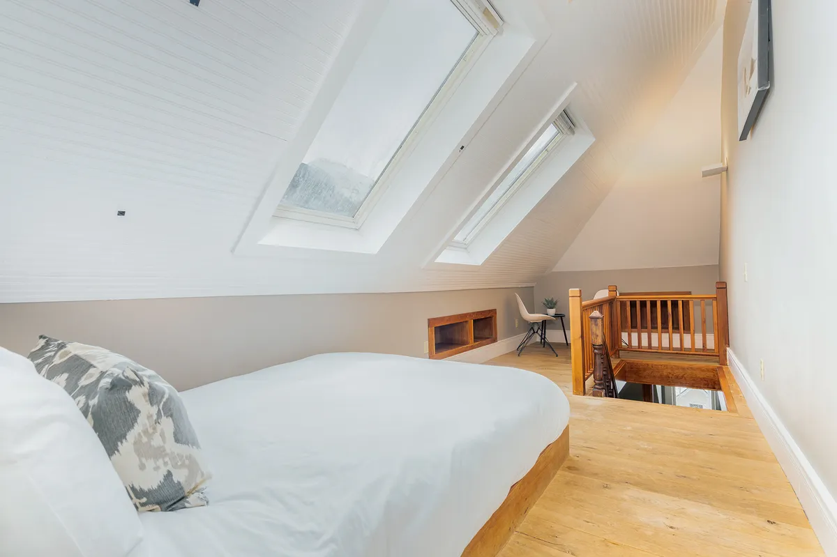 A cozy loft-style sleeping area with a neatly made bed beneath skylights, a minimalist chair, and a wooden railing. The soft, natural light creates an intimate ambiance, making the space perfect for children or close friends.
