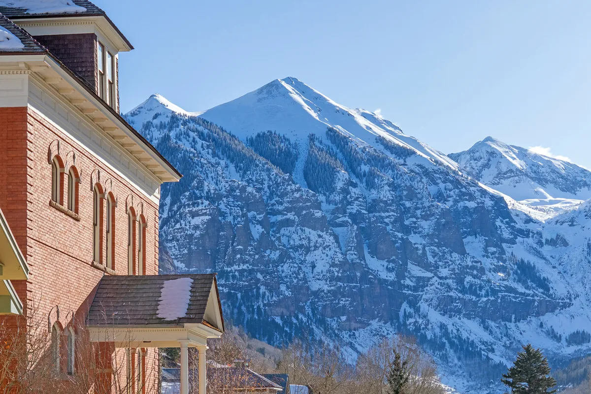 A historic brick building stands at the edge of downtown Telluride, with a striking snow-capped mountain backdrop. The sidewalk and surrounding area highlight proximity to both the town’s amenities and the great outdoors, setting the stage for adventure in every season.