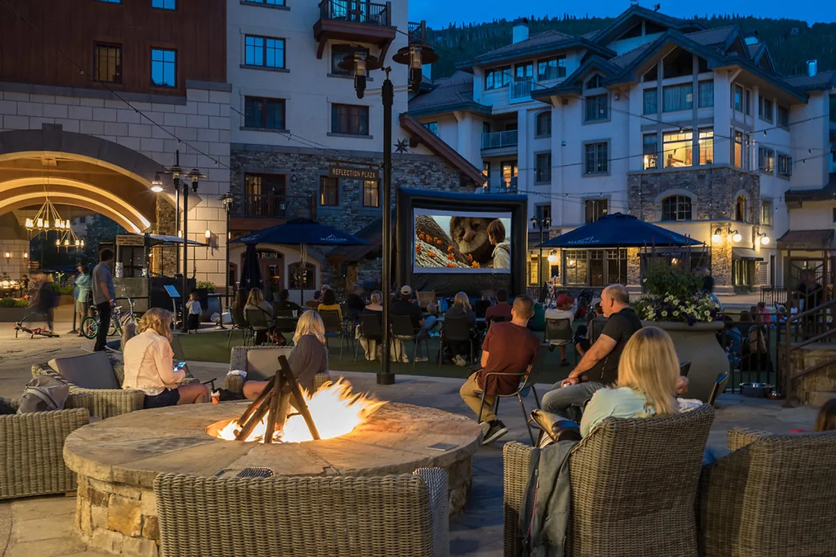 An inviting outdoor communal area featuring a fire pit, movie screen, and comfortable seating surrounded by natural scenery. The space is designed for socializing, relaxation, and making memories with friends or fellow travelers.