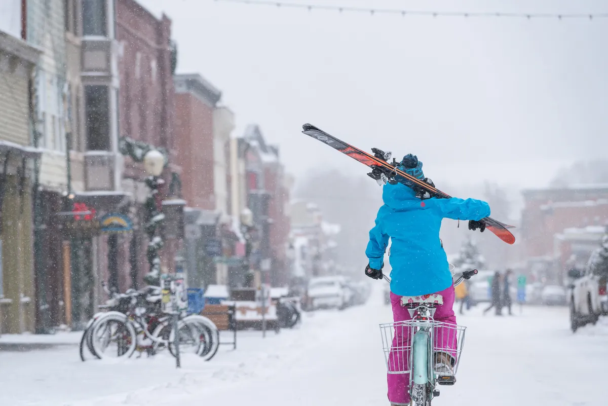 A charming street scene in winter, featuring historic buildings with a dusting of snow. A person in the foreground pedals a bicycle while carrying skis, with parked bikes and lively shopfronts visible. The atmosphere is playful and hints at Telluride's unique blend of heritage and active lifestyle.