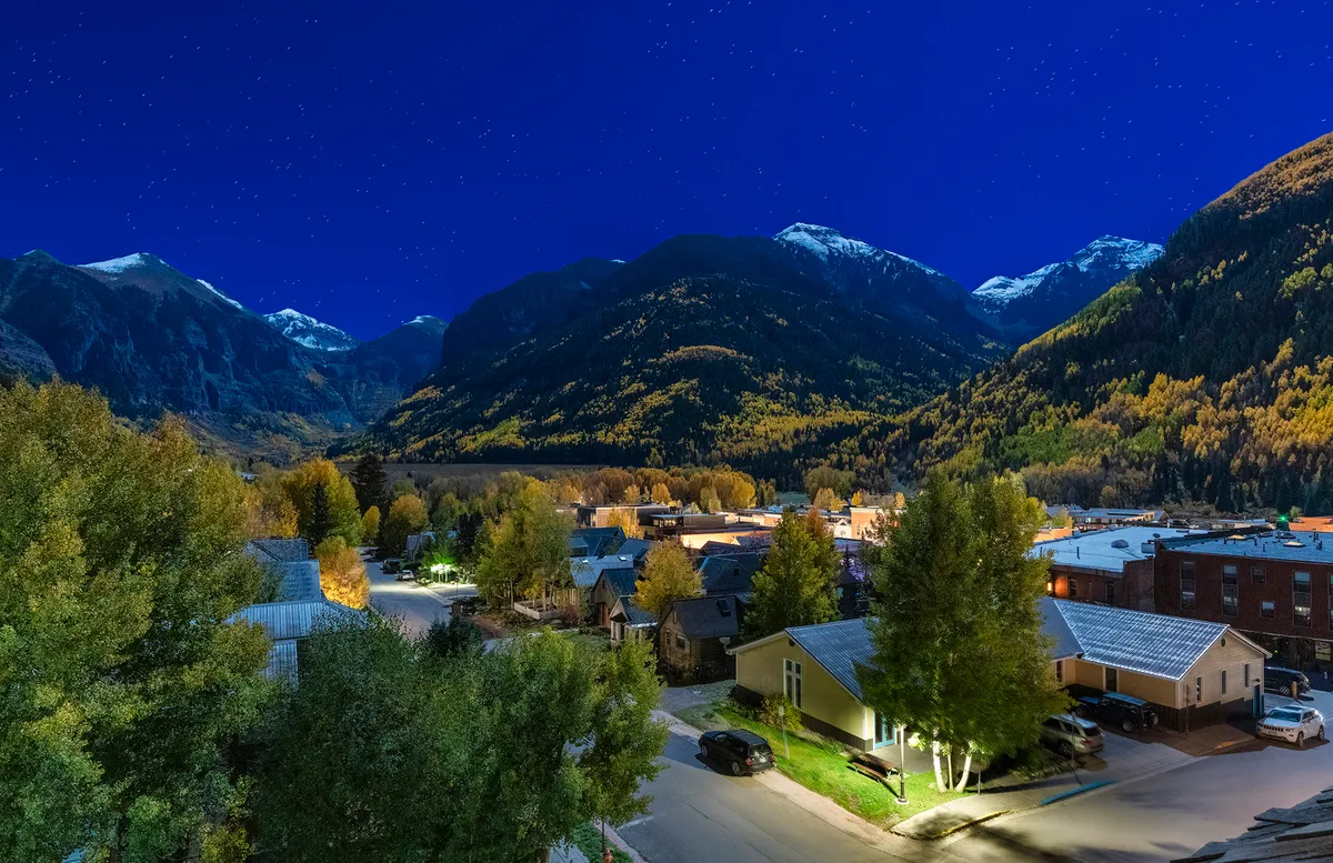 A panoramic nighttime view of Telluride reveals mountains silhouetted against the sky, twinkling stars, and glowing streetlights illuminating the town below. The scene is calm and inviting, evoking a sense of adventure and relaxation in the evening hours.