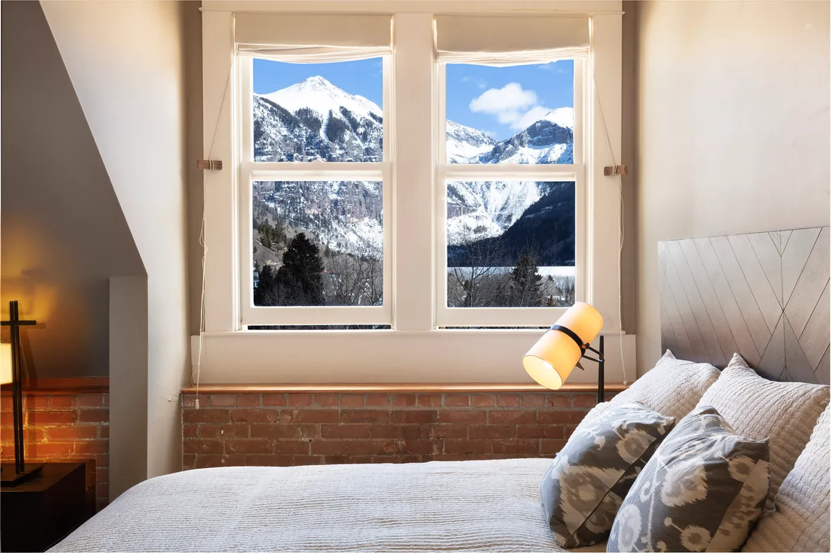 A sunlit, cozy loft bedroom with a plush bed positioned near large windows, offering breathtaking, unobstructed views of Ajax mountain. The room features soft lighting from a bedside lamp, inviting guests to relax, read, or simply soak in the mountain scenery right from bed. The historic character of exposed brick and original architecture is visible, creating a warm, inviting atmosphere.
