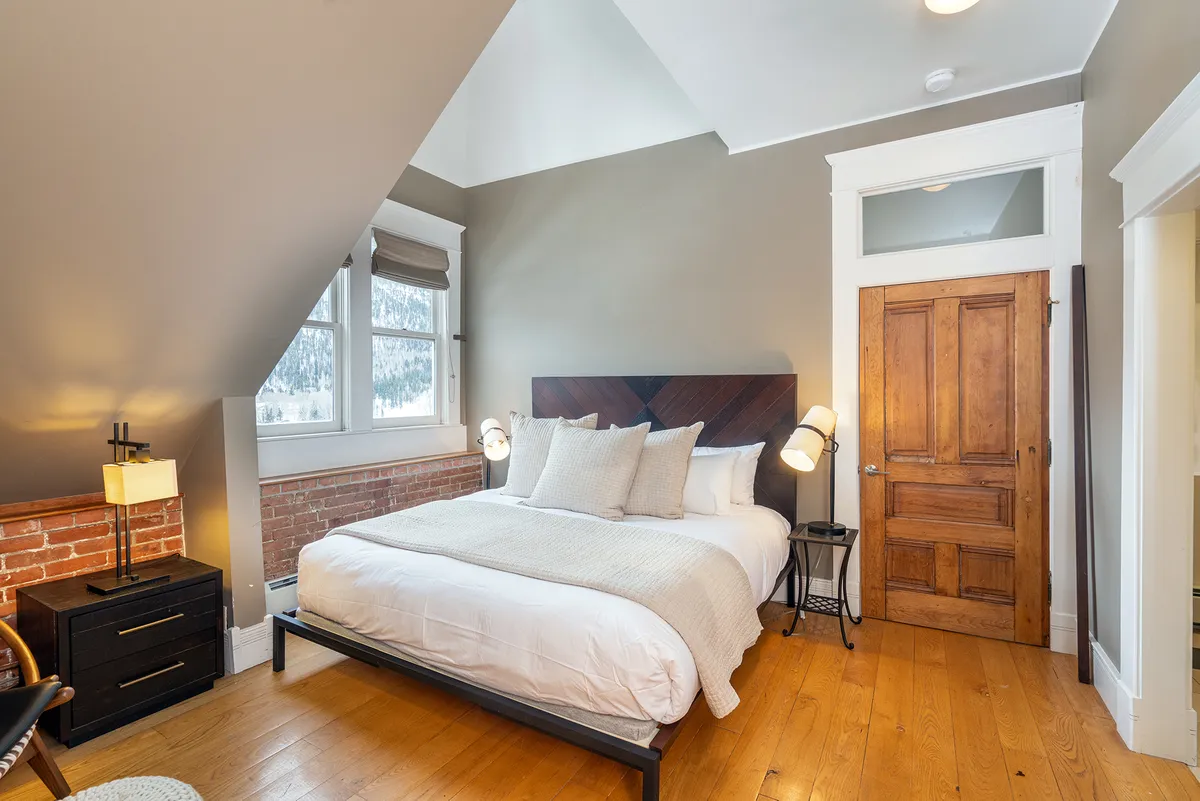 A bright, relaxed bedroom featuring a comfortable bed dressed in soft linens, a large window allowing natural light to pour in, a rustic brick accent wall, and a wooden door. The space exudes tranquility and comfort—ideal for restful nights or rejuvenating mornings with mountain views.