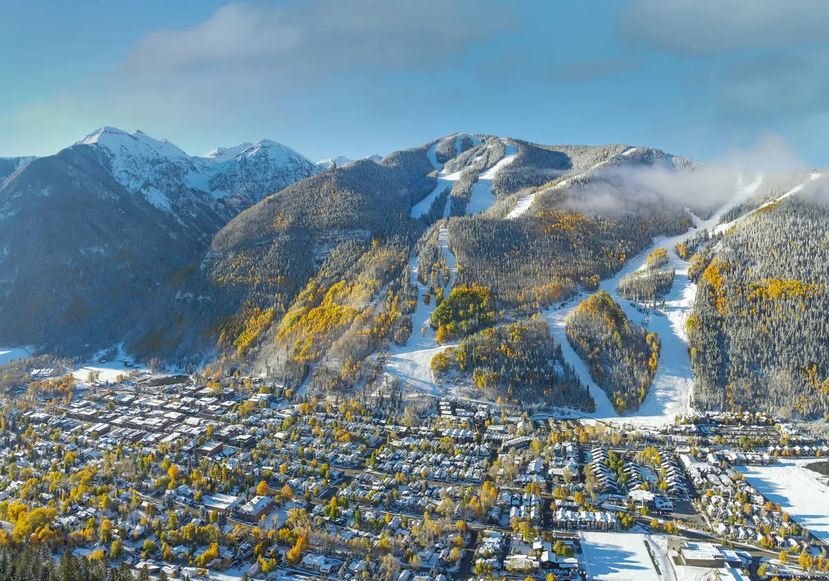 A sweeping view from above of Telluride, showing snow-capped mountains, ski slopes, autumn-colored trees, and the quaint town nestled below. The scene captures the natural beauty and unique mountain setting that guests can explore during their stay.