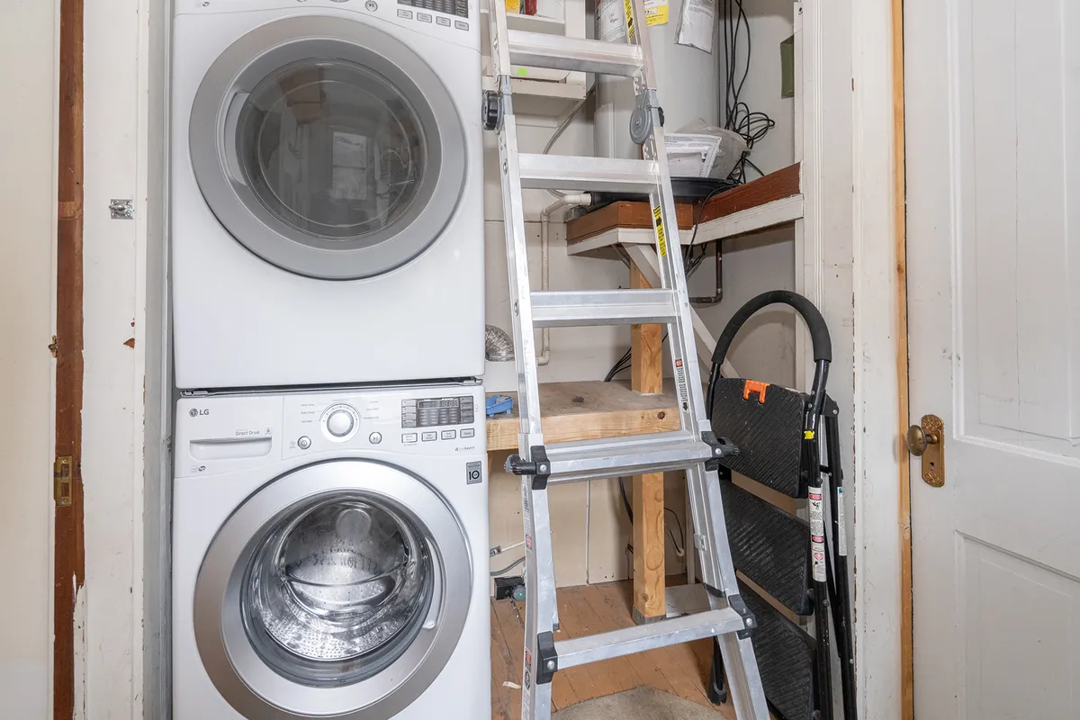A practical, organized laundry area is depicted with a stacked washer and dryer, step ladder, and shelves filled with supplies. The mood is functional and clean, emphasizing convenience and utility for longer stays or active guests.