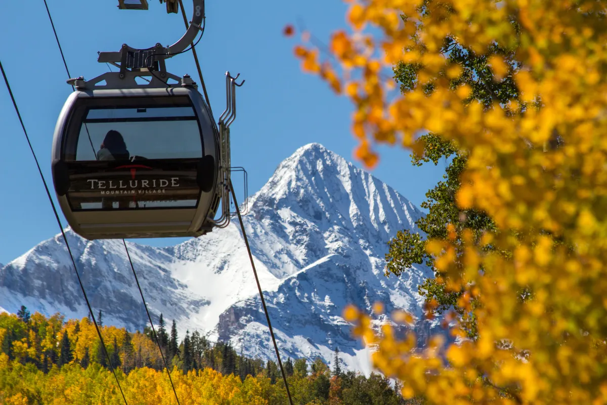 A gondola car glides over autumn trees toward a snow-capped mountain, perfectly encapsulating Telluride’s year-round outdoor appeal. The view blends vibrant fall foliage, snowy peaks, and the iconic gondola, hinting at hiking, skiing, and scenic rides.