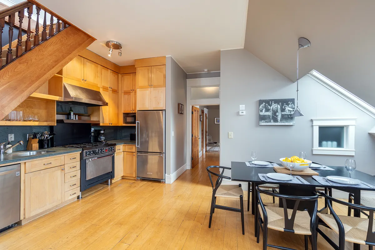 An inviting, open-plan kitchen and dining space where shiny appliances, a central dining table, and a staircase are visible. The layout encourages conversation and togetherness while cooking or dining.