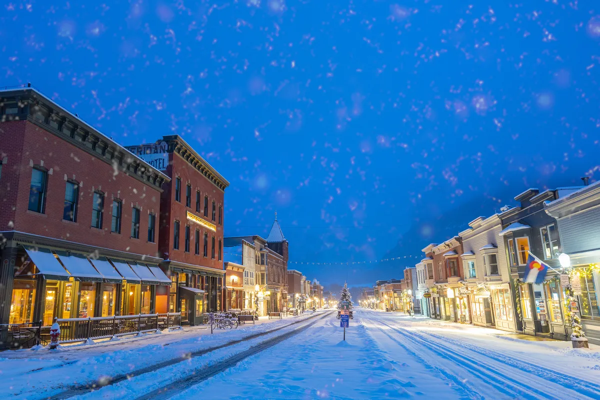 A picturesque winter street scene in downtown Telluride, with snow-covered roads, festive storefronts, and a distant Christmas tree, evoking the town's vibrant atmosphere during seasonal events.