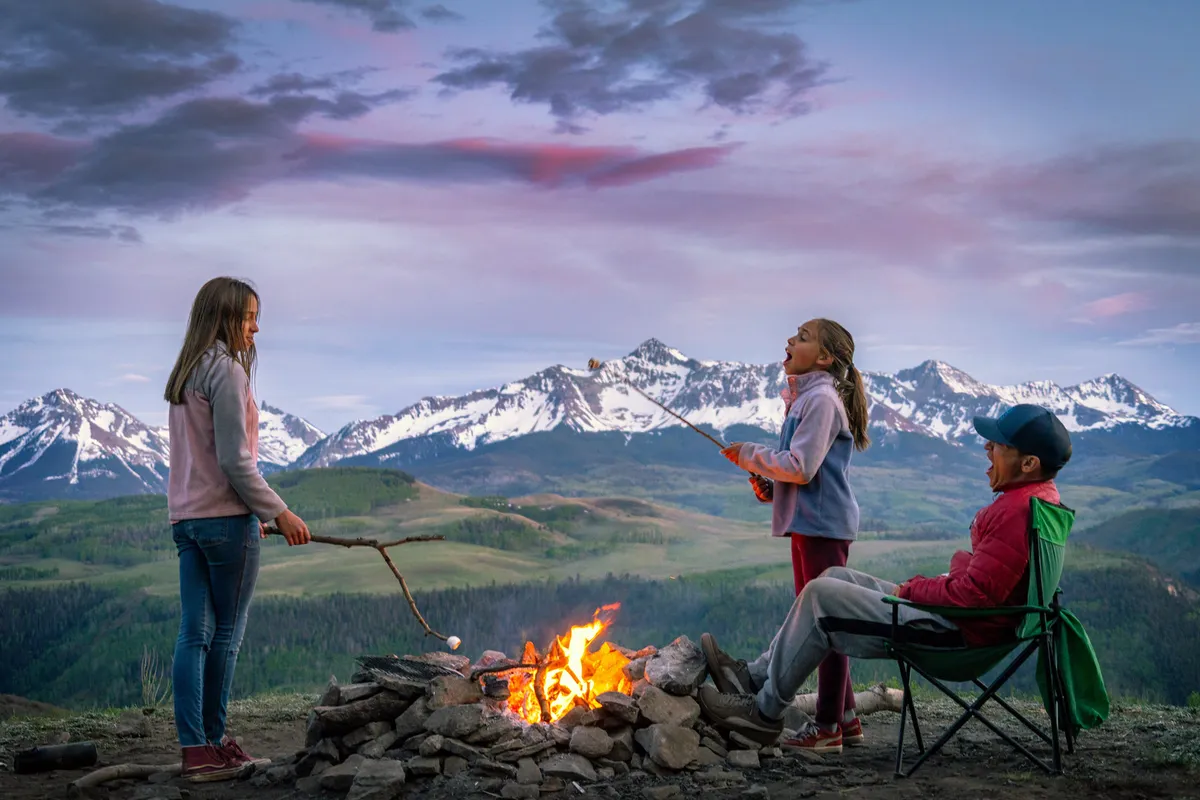 Children roast marshmallows around a campfire as dusk settles over the mountains. A man relaxes in a chair, creating a scene of family togetherness and outdoor fun, with nature’s beauty ever-present in the background.