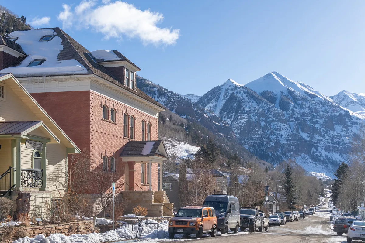 A prominent brick building stands at the heart of Telluride, framed by a backdrop of snow-covered mountains and a tree-lined street. The scene suggests a lively, walkable neighborhood filled with local charm and a sense of welcoming community.