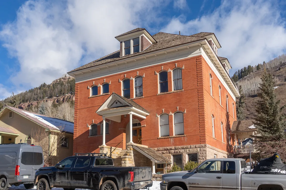 This image captures the exterior of the Miner's Union building, showcasing its historic red brick facade, large windows, and the surrounding mountain landscape. The inviting front entrance hints at easy access to the vibrant town beyond.